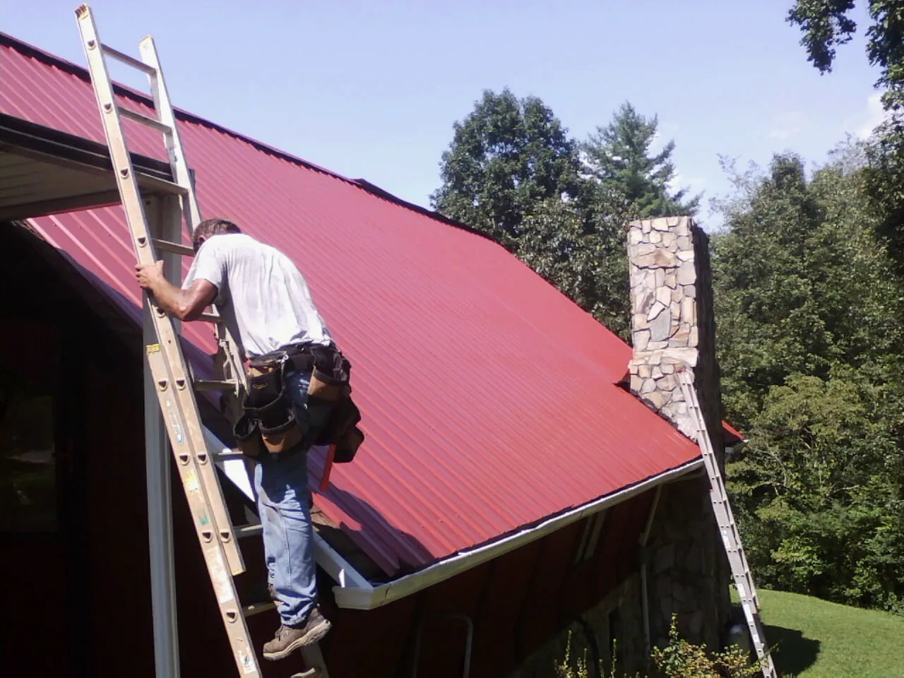 Roofer on a ladder beside a red metal roof and stone chimney — Soffit & Fascia Repair by Roofing Brothers in Powhatan Point